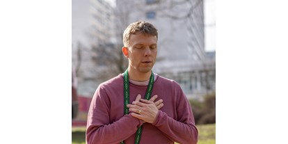 Yogakurs - Ambiente: Modern - Hamburg-Stadt Farmsen - Ein Mann in meditativer Pose mit geschlossenen Augen und verschränkten Händen über dem Herzen, gekleidet in einen lila Pullover und mit einem grünen Schal um den Hals, der die Ruhe und Zentrierung einer Yin Yoga-Praxis ausstrahlt. Der Hintergrund mit unscharfen Gebäuden suggeriert, dass die Praxis in einer städtischen Umgebung stattfindet. - SOMATiC YiNYOGA